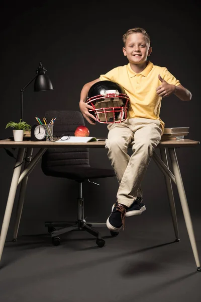 Очаровательный школьник держит американский футбольный шлем, делает большой палец вверх жестом и сидит на столе с книгами, растением, очками, лампой, цветными карандашами, яблоком, часами и учебником на сером фоне — стоковое фото
