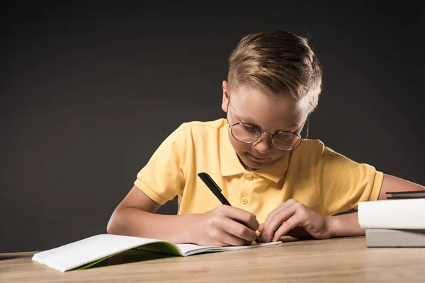 Colegial enfocado en anteojos escribiendo en libro de texto en la mesa con pila de libros sobre fondo gris - foto de stock