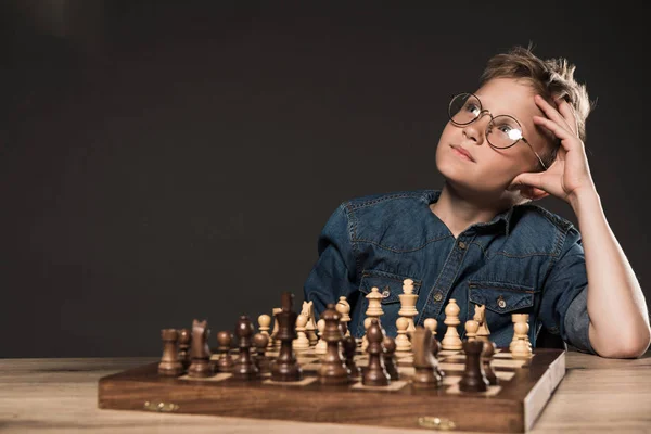 Petit garçon réfléchi dans des lunettes assis à table avec échiquier sur fond gris — Photo de stock