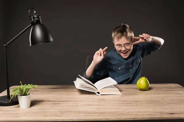 Menino animado em óculos fazendo sinais de paz à mesa com livro, pêra, planta e lâmpada em fundo cinza — Fotografia de Stock