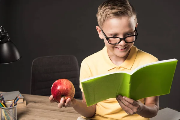 Écolier souriant en lunettes tenant la pomme et faisant ses devoirs près de la table avec crayons de couleur, lampe et livres sur fond gris — Photo de stock