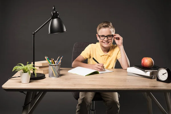 Piccolo scolaretto in occhiali che fa i compiti a tavola con libri, piante, lampade, matite a colori, mela, orologio e libro di testo su sfondo grigio — Foto stock