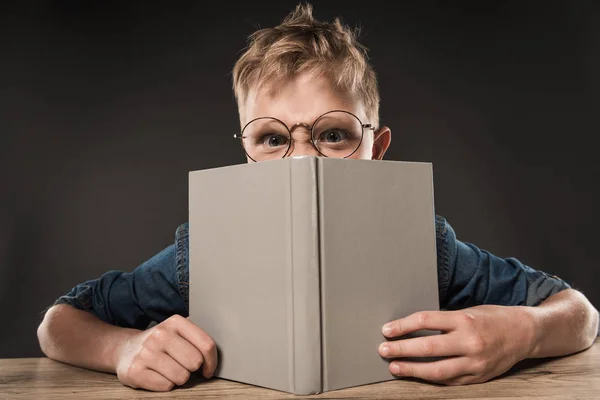 Colegial enojado en anteojos cubriendo cara por libro en la mesa sobre fondo gris - foto de stock