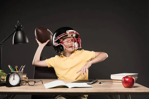 Школьник в американском футбольном шлеме бросает мяч и сидит за столом с книгами, растением, лампой, цветными карандашами, яблоком, часами и учебником на сером фоне — стоковое фото