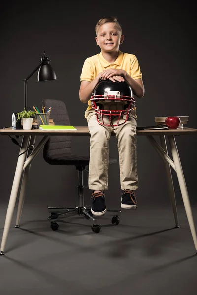 Colegial sonriente sosteniendo casco de fútbol americano y sentado en la mesa con libros, planta, lámpara, lápices de color, manzana, reloj y libro de texto sobre fondo gris - foto de stock