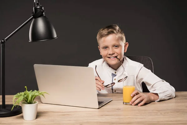 Petit garçon souriant tenant des lunettes et assis à table avec un verre de jus, ordinateur portable, plante et lampe sur fond gris — Photo de stock