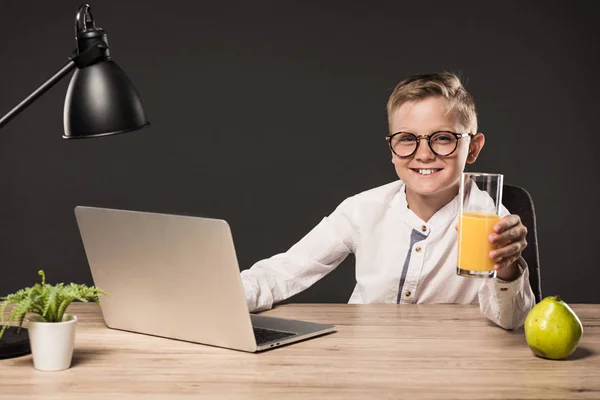 Niño feliz en gafas con vaso de jugo y sentado en la mesa con el ordenador portátil, la planta y la lámpara sobre fondo gris - foto de stock