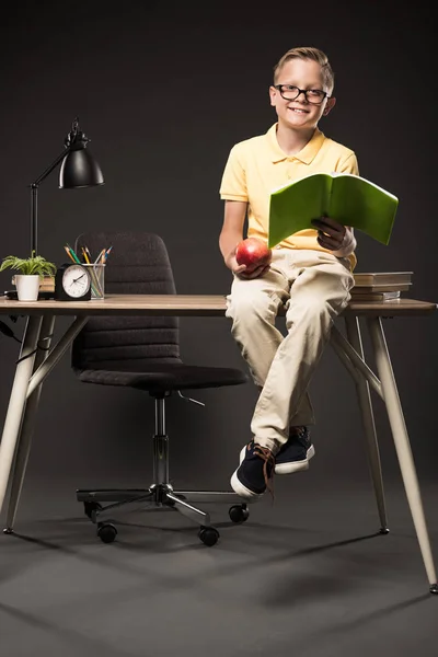 Colegial sonriente sosteniendo manzana y haciendo los deberes mientras está sentado en la mesa con libros, plantas, lámparas, lápices de color, reloj y libro de texto sobre fondo gris - foto de stock