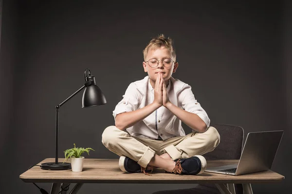 Мальчик с закрытыми глазами в очках медитирует и сидит на столе с ноутбуком, растением и лампой на сером фоне — стоковое фото