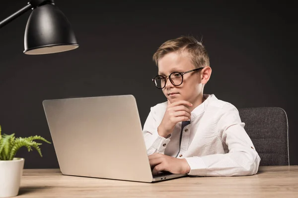 Serious little boy in eyeglasses using laptop at table with plant and lamp on grey background — Stock Photo
