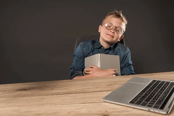 Little schoolboy in eyeglasses sleeping with book at table with laptop on grey background — Stock Photo