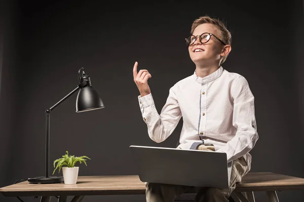 Улыбающийся маленький мальчик в очках делает идею жестом за пальцем и с помощью ноутбука, сидя на столе с лампой и растением на сером фоне — стоковое фото