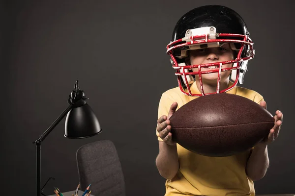 Niño feliz en casco de fútbol americano sosteniendo la pelota sobre fondo gris - foto de stock