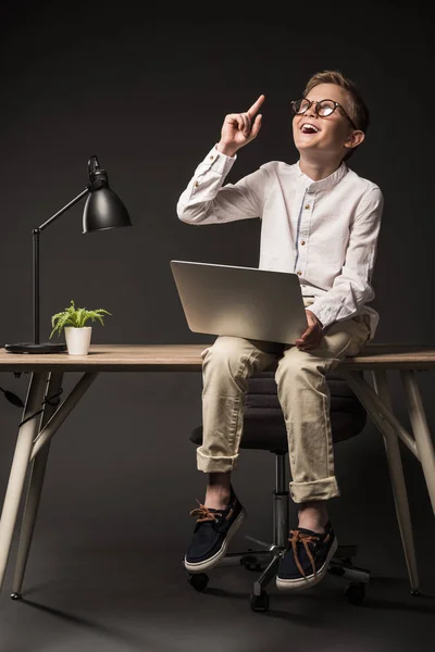 Возбужденный маленький мальчик в очках делает идею жест за пальцем и с помощью ноутбука, сидя на столе с лампой и растения на сером фоне — стоковое фото