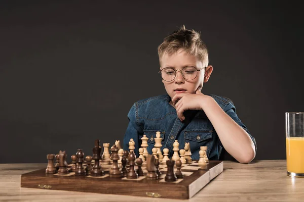 Niño pensativo sentado en la mesa con tablero de ajedrez y vaso de jugo sobre fondo gris — Stock Photo