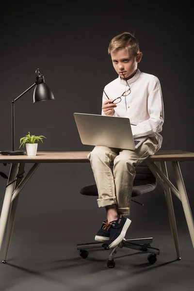 Menino grave segurando óculos e usando laptop enquanto sentado na mesa com planta e lâmpada no fundo cinza — Fotografia de Stock