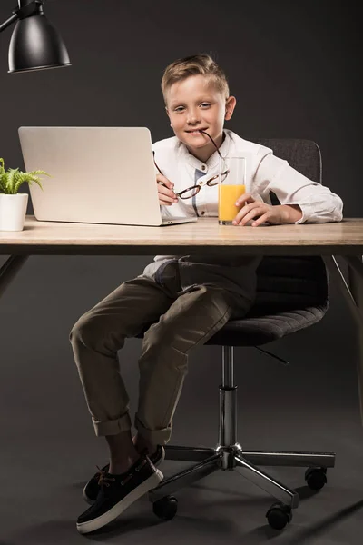 Petit garçon souriant tenant des lunettes et assis à table avec ordinateur portable, verre de jus, plante et lampe sur fond gris — Photo de stock