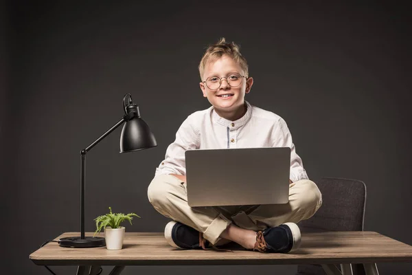 Niño sonriente en gafas usando portátil mientras está sentado en la mesa con la planta y la lámpara sobre fondo gris - foto de stock