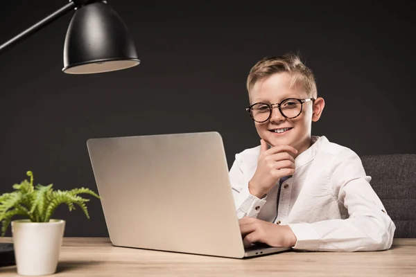 Bambino sorridente con gli occhiali che tiene la mano sul mento e siede a tavola con computer portatile, pianta e lampada su sfondo grigio — Foto stock