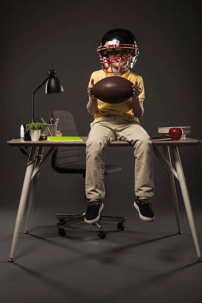 Colegial sonriente en casco de fútbol americano sosteniendo pelota y sentado en la mesa con libros, planta, lámpara, lápices de color, manzana, reloj y libro de texto sobre fondo gris - foto de stock