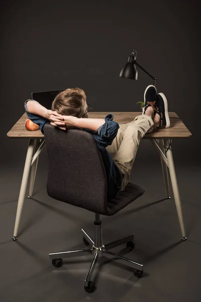 Vista trasera del niño descansando con las piernas en la mesa con el ordenador portátil, manzana y lámpara sobre fondo gris - foto de stock