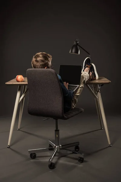 Вид сзади маленького мальчика с помощью ноутбука и сидя с ногами на столе на сером фоне — стоковое фото