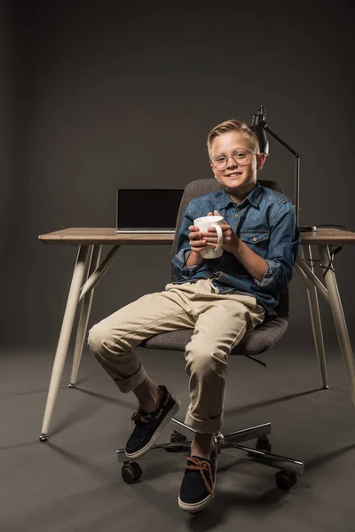 Little boy in eyeglasses holding coffee cup and sitting on chair near table with laptop and lamp on grey background — Stock Photo