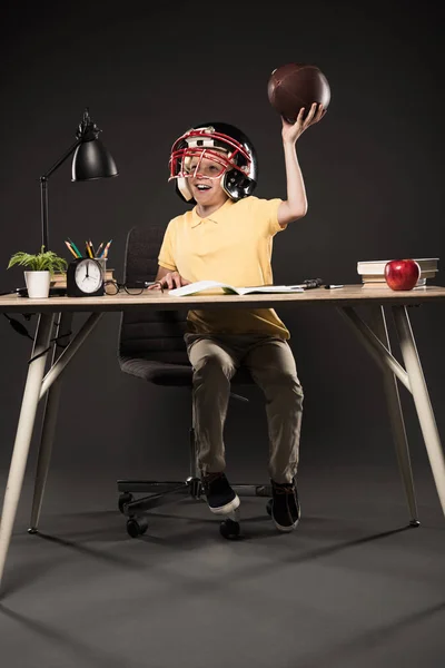 Счастливый школьник в американском футбольном шлеме держит мяч и сидит за столом с книгами, растением, лампой, цветными карандашами, яблоком, часами и учебником на сером фоне — стоковое фото