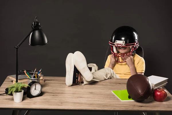 Улыбающийся школьник в американском футбольном шлеме сидит с ногами на столе с мячом, книгами, растением, лампой, цветными карандашами, яблоком, часами и учебником на сером фоне — стоковое фото