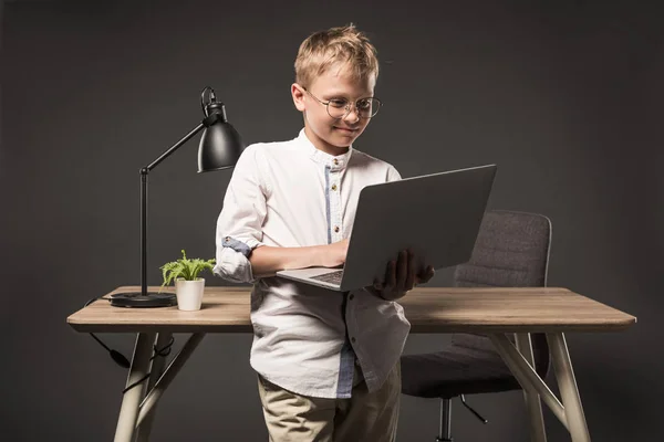 Niño pequeño en gafas con portátil cerca de la mesa con la planta y la lámpara sobre fondo gris - foto de stock