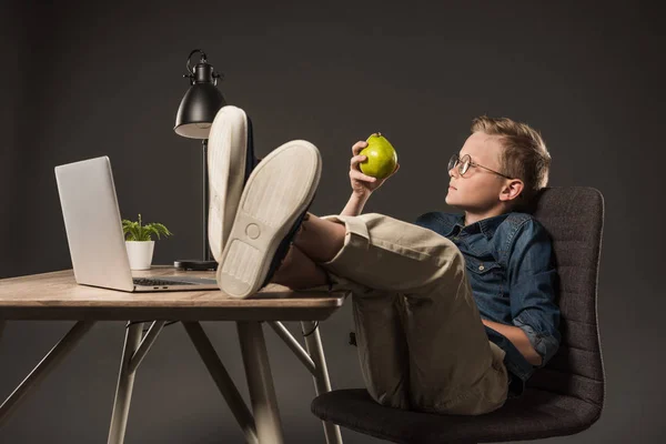 Vista laterale del bambino in occhiali che tiene la pera e seduto con le gambe sul tavolo con laptop, pianta e lampada su sfondo grigio — Foto stock