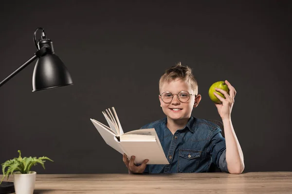 Estudante sorrindo segurando pêra e lendo livro à mesa com planta e lâmpada em fundo cinza — Fotografia de Stock