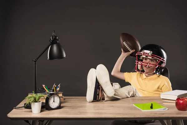 Colegial sonriente en casco de fútbol americano sosteniendo pelota y sentado con las piernas en la mesa con libros, planta, lámpara, lápices de color, manzana, reloj y libro de texto sobre fondo gris - foto de stock
