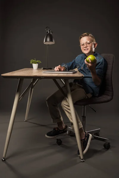 Smiling little boy in eyeglasses holding pear and sitting at table with laptop, lamp and plant on grey background — Stock Photo