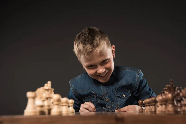 Menino feliz segurando óculos e olhando para tabuleiro de xadrez na mesa isolado em fundo cinza — Fotografia de Stock