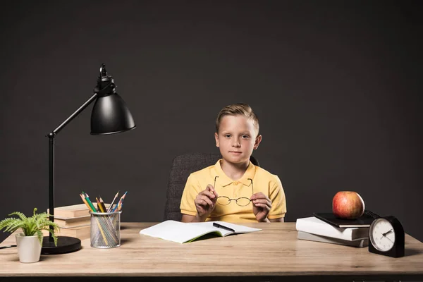 Schoolboy holding eyeglasses and sitting at table with books, plant, lamp, colour pencils, apple, clock and textbook on grey background — Stock Photo