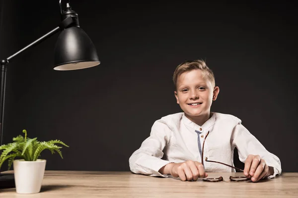 Niño feliz sentado a la mesa con anteojos, planta y lámpara sobre fondo gris - foto de stock