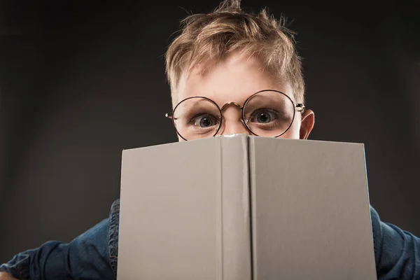 Little schoolboy in eyeglasses covering face by book isolated on grey background — Stock Photo