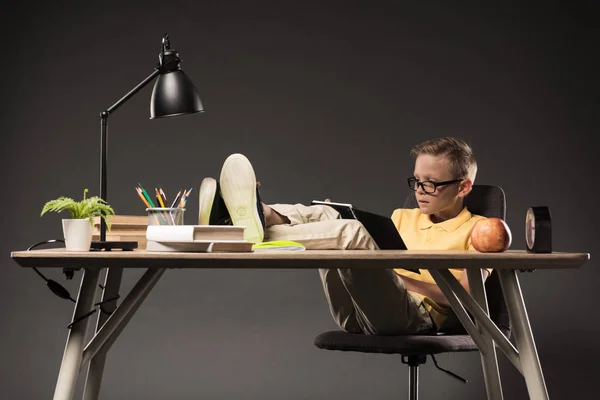 Colegial en gafas haciendo la tarea, sosteniendo manzana y sentado con las piernas en los libros de mesa, planta, lámpara, lápices de color, reloj y libro de texto sobre fondo gris - foto de stock