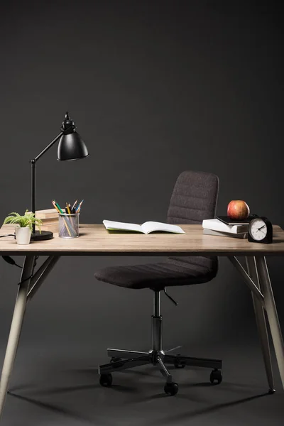 Vorderseite des Tisches mit Lampe, Pflanze, Büchern, Farbstiften, Brillen, leerem Lehrbuch, Apfel und Uhr auf grauem Hintergrund — Stockfoto