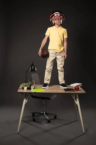 Школярка в американському футбольному шоломі тримає м'яч і стоїть на столі з книгами, рослинами, лампами, кольоровими олівцями, яблуком, годинником і підручником на сірому фоні — стокове фото
