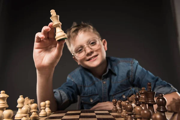 Selective focus of little boy in eyeglasses holding chess figure over chess board on grey background — Stock Photo