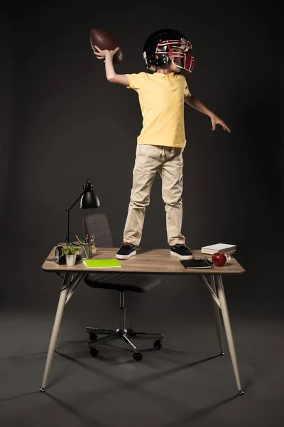 Школяр в американському футбольному шоломі кидає м'яч і стоїть на столі з книгами, рослинами, лампами, кольоровими олівцями, яблуком, годинником і підручником на сірому фоні — стокове фото