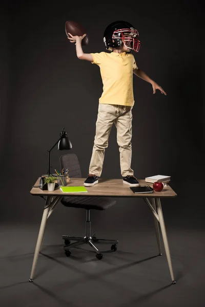 Schoolboy in american football helmet throwing ball and standing on table with books, plant, lamp, colour pencils, apple, clock and textbook on grey background — Stock Photo