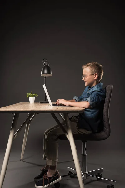 Side view of happy schoolboy using laptop at table with lamp and potted plant on grey background — Stock Photo