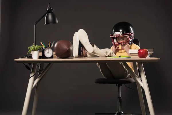 Colegial sonriente en casco de fútbol americano sentado con las piernas en la mesa con bola, libros, planta, lámpara, lápices de color, manzana, reloj y libro de texto sobre fondo gris - foto de stock