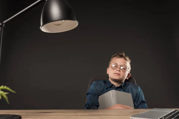 Petit écolier en lunettes dormant avec livre dans les mains à table sur fond gris — Photo de stock