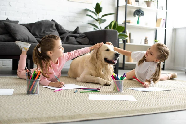 Adorable kids petting golden retriever dog while drawing pictures on floor at home — Stock Photo