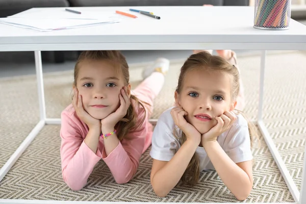 Retrato de hermanas pequeñas mirando a la cámara mientras yacen debajo de la mesa con lápices en casa - foto de stock