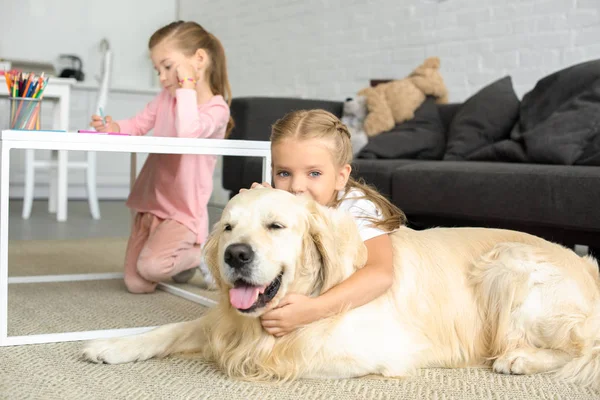 Selective focus of kid hugging golden retriever dog on floor while sister drawing picture at home — Stock Photo